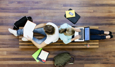 Eine Frau mit Laptop und ein Mann mit Buch sitzen Rücken an Rücken auf dem Boden. 