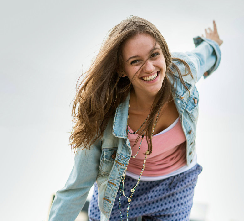 Eine junge Frau mit Jeansjacke lacht und breitet die Arme aus.  